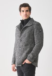 Hombre que lleva un abrigo abotonado de patrón de espiga en gris sobre una chaqueta negra con cremallera, con una mano en el bolsillo frente a un fondo blanco liso.