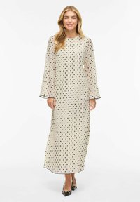 Lang beige kjole med svarte polkadotter, lange klokkeærmer og rett passform. Gjennomsiktig tekstur med svart kant ved halslinningen og mansjettene.
