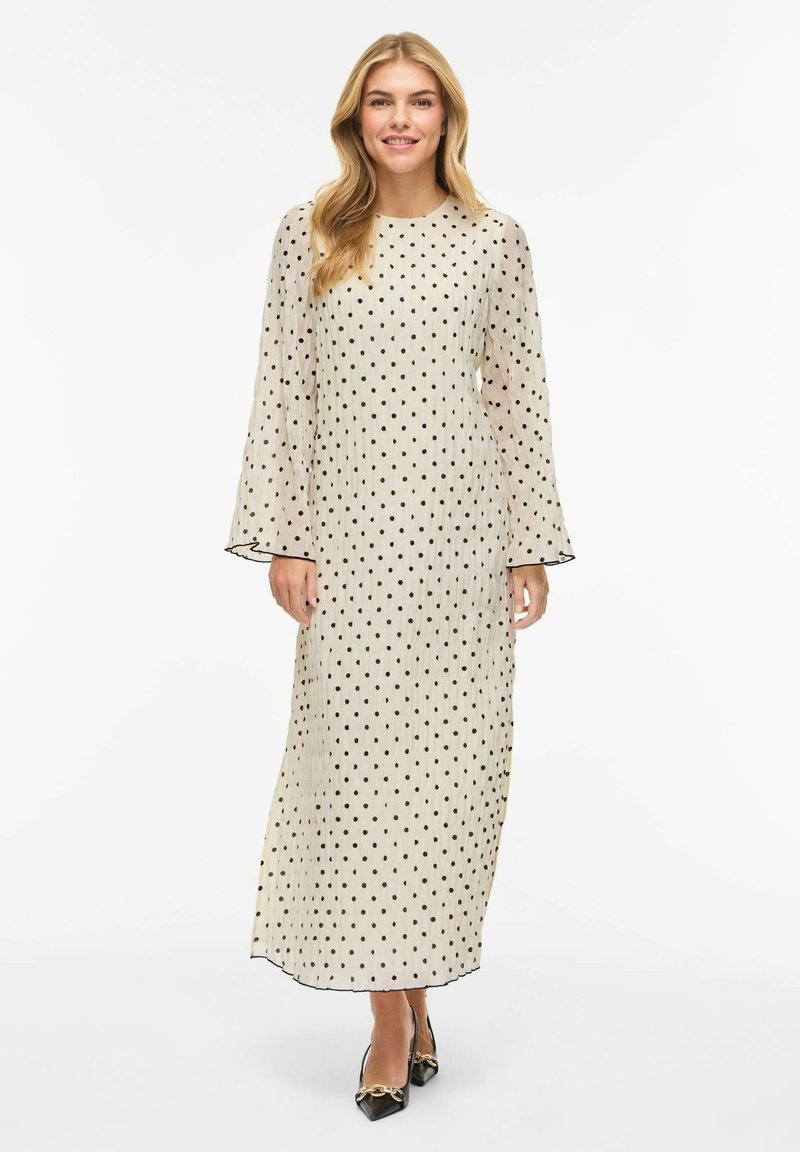 Lang beige kjole med svarte polkadotter, lange klokkeærmer og rett passform. Gjennomsiktig tekstur med svart kant ved halslinningen og mansjettene.