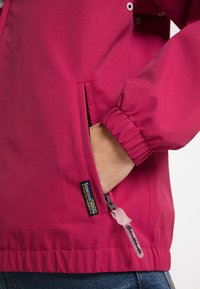 Fuchsia jacka tillverkad av lätt tyg, med elastiska manschetter, en ficka med dragkedja och en logotags på sidan.