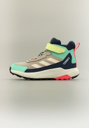 Vysoký teniska Adidas Terrex v béžovej, mätovo zelenej, námorníckej modrej a neónovo žltej farbe s pásikom na suchý zips a červenou pútkom na päte na svetlom pozadí.