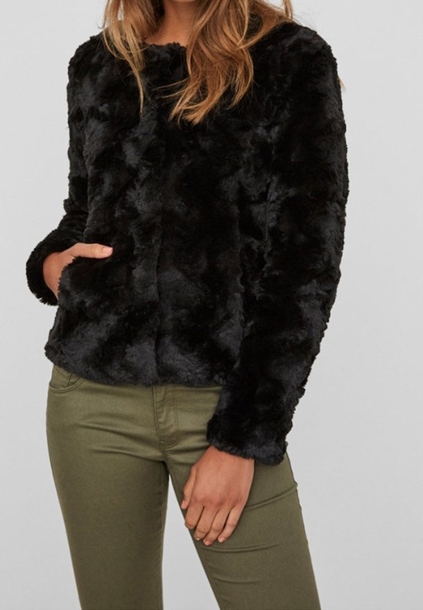 Femme portant une veste en fausse fourrure noire à manches longues et un pantalon vert ajusté sur un fond uni.