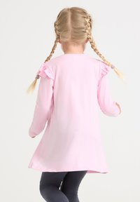 Tunica rosa a maniche lunghe con dettagli arricciati sulle spalle, realizzata in tessuto morbido. Abbinata a leggings scuri, presenta un orlo leggermente curvo.