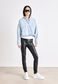 Lichtblauwe denim pullover met een halve rits, zwarte nepleer leggings en witte platform sneakers; zwarte zonnebril maakt de look compleet.