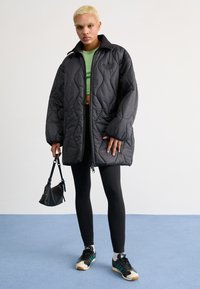 Padded melnā jaka ar kvadrātu intensīvumu, augsts apkakles un rāvējslēdzis. Uzvilkta virs zaļā topa, kombinēta ar melnām legingām un sporta apaviem.