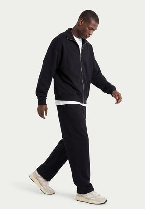 Homme marchant, portant une veste noire à fermeture éclair, un pantalon noir, un sous-vêtement blanc et des baskets beiges, sur un fond clair uni.