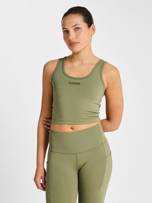 Kvinde iført olivengrøn ærmeløs croptop og højtailede leggings, stående med den ene hånd bag ryggen mod en hvid baggrund.