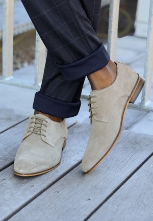 Beige nubukcipő, hegyes orral és fűzős dizájnnal. Fafurnérral ellátott talp, összepárosítva sötétkék kockás nadrággal, amelynek szárát feltekerték.