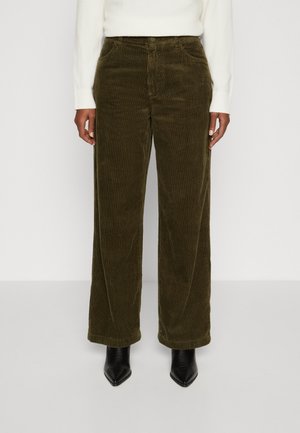 Person, die olivgrüne, weite Cordhosen, ein weißes langärmeliges Oberteil und schwarze spitze Stiefeletten trägt, steht vor einem schlichten Hintergrund.