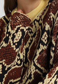 Strickpullover mit einem braunen, schwarzen und cremefarbenen Schlangenmotiv. Er hat einen gelben gerippten Kragen und lange Ärmel mit gefütterten Enden.