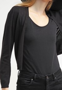 Kvinna som bär en svart topp med rundad halsringning, en kort svart cardigan och högmidjade svarta jeans mot en enfärgad bakgrund.