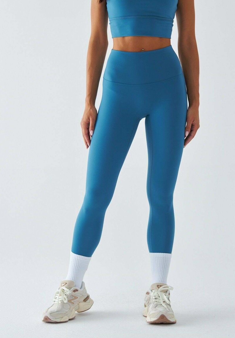 Zestaw aktywny w kolorze zielonym morskim, składający się z krótkawego topu i legginsów z wysokim stanem, oba o gładkiej teksturze; do tego białe krótkie skarpetki i jasne trampki.