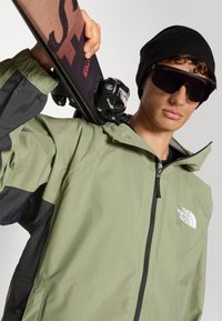 Grüne und schwarze Jacke mit Reißverschluss und Kapuze, getragen mit einer schwarzen Mütze und übergroßen Sonnenbrillen, hält einen Ski mit Bindungen.