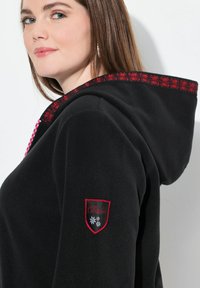 Hoodie en polaire noire avec un accent rouge brodé sur la capuche, présentant un patch logo sur la manche. Texture lisse et design ajusté.