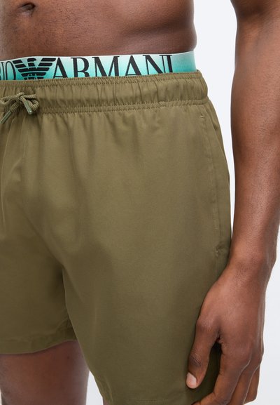 Ανδρικός κορμός που φοράει χακί σορτσάκι με κορδόνι και τιρκουάζ ζώνη Armani ορατή πάνω από το σορτς, με το δεξί χέρι χαλαρό στο πλάι.