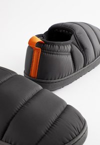 Chaussons noirs rembourrés avec semelle texturée et languette orange. Matériau extérieur doux et matelassé, design à enfiler pour un port facile.