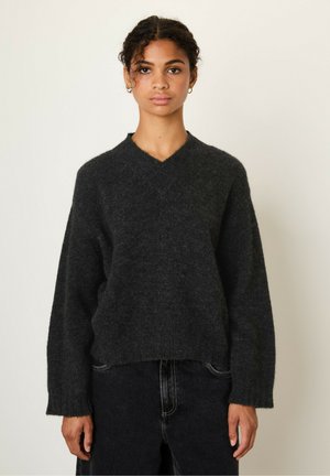 Young woman with curly hair wearing a dark gray fuzzy V-neck sweater and black pants, standing against a plain light background.