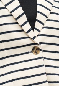 Blazer crème à rayures horizontales bleu marine, fermeture à un bouton en écaille de tortue, et chemise intérieure noire visible au col.