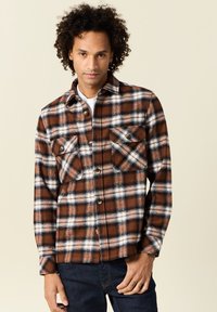 Giacca in flanella a quadri marrone e bianca con due tasche sul petto, chiusura a bottoni e colletto. Indossata sopra una maglietta bianca e jeans di denim.