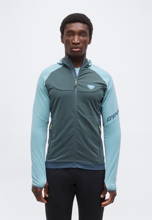 Muž, ktorý má na sebe tyrkysovo-šedú športovú bundu so zipsom a otvorom na palce a čierne nohavice, stojaci na jednoliatom bielom pozadí.