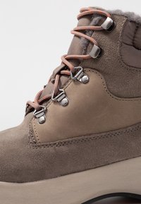 Brune ruskindsankelstøvle med grå tekstiloverdel, der har metalhooks og orange snørebånd. Markante teksturerede detaljer og gummisål.