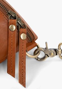 Braune Ledertasche Ecke mit bronzefarbenem Reißverschluss und Beschlägen; verfügt über zwei Lederriemen mit Metallnieten, detaillierter Naht und einem Clip-Anhänger.