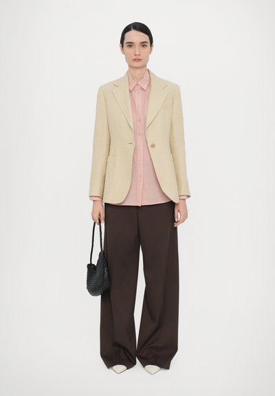 Beige blazer over a pink shirt, brown wide-leg trousers, and white pointed shoes. Holding a black woven bag. Simple design and modern fit.
