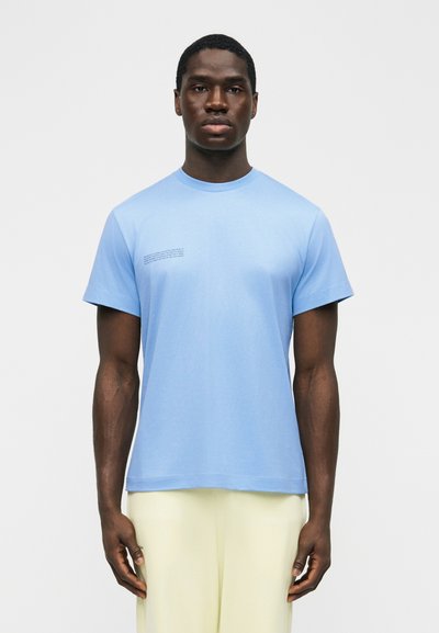 Άνδρας που φοράει ανοιχτό γαλάζιο κοντομάνικο μπλουζάκι με μικρό κείμενο στο αριστερό στήθος και ανοιχτό μπεζ παντελόνι, στέκεται μπροστά σε απλό λευκό φόντο.