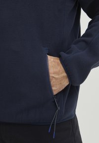 Dunkelblaues Fleece-Pullover mit einer Reißverschlusstasche, elastischen Bündchen und einer weichen Textur. Die Reißverschluss-Zipper sind in Blau akzentuiert.