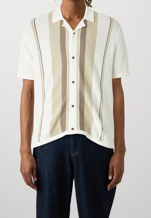 Mann trägt ein kurzärmeliges Strick-Poloshirt mit vertikalen beige-weißen Strukturstreifen, durchgehend geknöpft, kombiniert mit dunkelblauer Jeans.