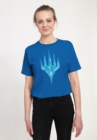 Modrá bavlněná tričko s geometrickým, ledovým designem ruky v odstínech světle modré. Krátké rukávy a kulatý výstřih. Nosené s černými kalhotami.