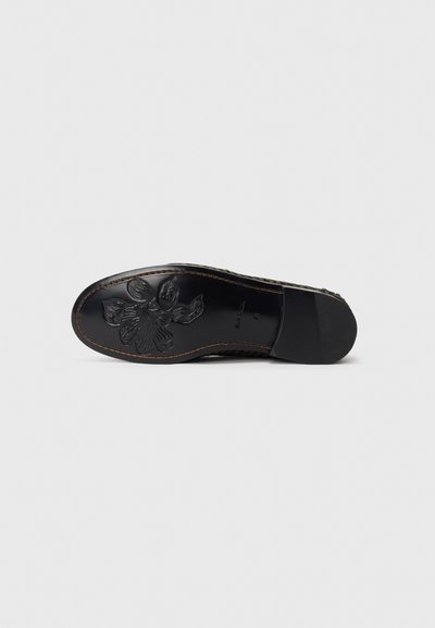 Suela de un zapato de cuero negro con costuras marrones y un patrón floral embossado cerca de la puntera, sobre un fondo plano.