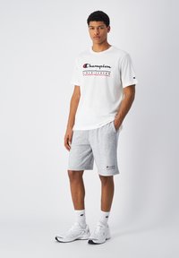 Camiseta de algodón blanca con el logo "Champion" en negro y rojo, combinada con pantalones cortos de algodón grises que presentan el logo de la marca, zapatillas deportivas blancas y calcetines blancos.