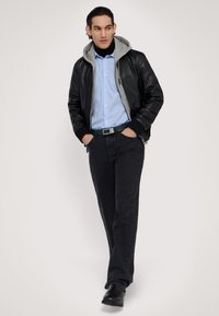 Černá kožená bomber bunda s šedou kapucí, modré pruhované tričko, zelený opasek, černé džíny a černé boty. Model má neutrální postoj.