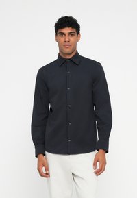 Camisa negra de manga larga con cuello puntiagudo, botonadura en la parte delantera y puños estándar, hecha de tela suave. Modelo vistiendo pantalones de color claro.