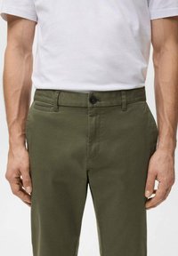 Mężczyzna w oliwkowozielonych spodniach i włożonej do środka gładkiej białej koszulce, ręce swobodnie opuszczone wzdłuż ciała, widoczne są talia i górna część nóg.