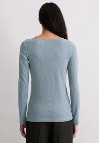 Gaiši zils garā piedurkne tops, izgatavots no mīksta, teksturēta auduma. Tam ir plaša, noapaļota kakla izgriezums un pieguļošs siluets.