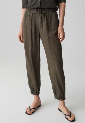 Donna che indossa pantaloni cargo verde oliva con coulisse e polsini elasticizzati e infradito nere su sfondo grigio chiaro.
