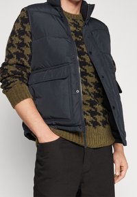 Vadderad svart quiltad väst med hög krage, som har två framfickor, bärs över en olivgrön mönstrad tröja och mörka byxor.