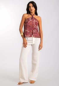 Haut à encolure licou dans un motif paisley rouge et rose, associé à un pantalon large blanc et des sandales plates. Il présente une fermeture nouée au cou.