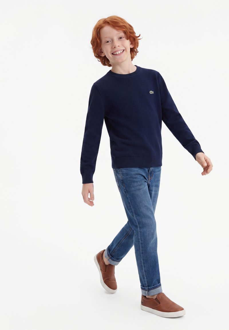 Jeune garçon roux souriant, portant un pull bleu marine, un jean bleu et des chaussures marron sans lacets, marchant sur fond blanc.