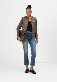 Braune gesteppte Bomberjacke mit grauen Ärmeln, marineblauem Oberteil und hellen weit geschnittenen Jeans. Schwarze Loafer und eine braune strukturierte Handtasche.