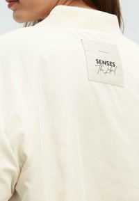Cream-colored bomber jacket featuring a ribbed collar and a label on the back with "SENSES The Label" in black text. Smooth texture.
