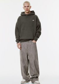 Felpa con cappuccio verde scuro, dotata di tasca frontale e logo bianco, abbinata a pantaloni grigi a righe verticali molto ampi e scarpe da ginnastica chiare.