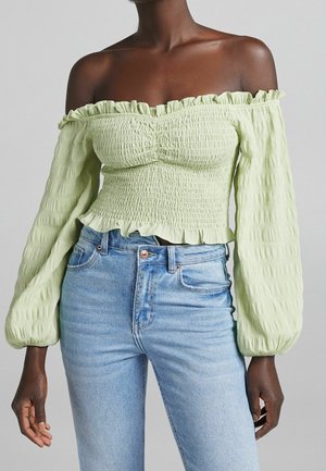 Femme portant un haut smocké vert clair à épaules dénudées avec de longues manches bouffantes et un jean taille haute bleu clair, debout sur un fond gris.