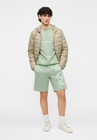 Geacă ușoară, matlasată, de culoare bej, set format din tricou verde și pantaloni scurți. Logo alb pe haine, pantofi sport asortați și șosete albe cu striații.