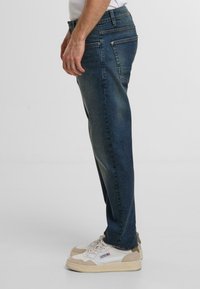 Jeans di denim blu scuro con una vestibilità dritta, dotati di tasche laterali e posteriori, una leggera scoloritura e cuciture a contrasto. Abbinati a sneakers di colore chiaro.