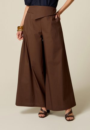 Wijde bruine broek met hoge taille en asymmetrische tailleband, gedragen met donkere sandalen en een marineblauwe top, gecombineerd met grove armbanden.