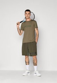 Olivengrønt treningsantrekk bestående av en kortermet skjorte med "BORG"-logo og matchende shorts, kombinert med hvite sportssko.
