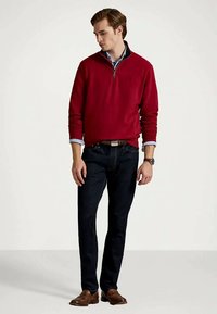 Młody mężczyzna w czerwonym swetrze z zamkiem błyskawicznym noszonym na niebieskiej koszuli, ciemnych dżinsach, brązowym pasku, zegarku i brązowych eleganckich butach, stojący na tle gładkiej ściany.